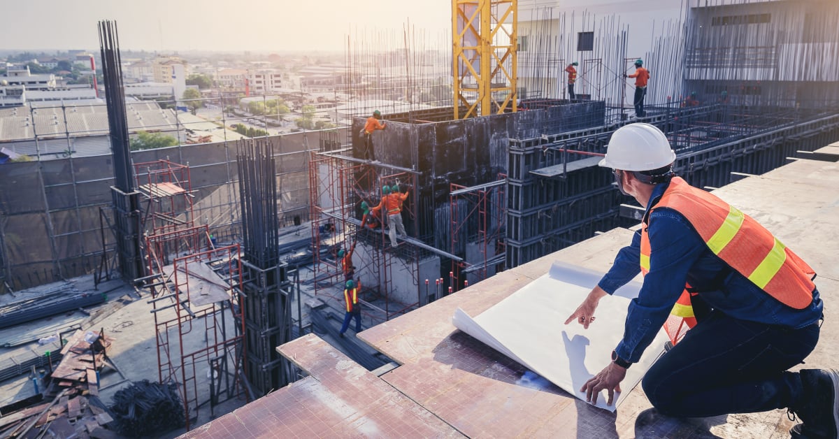 Construction supervisor in a white hard hat and orange safety vest kneels on a rooftop reviewing building plans while a crew works on the structure below.