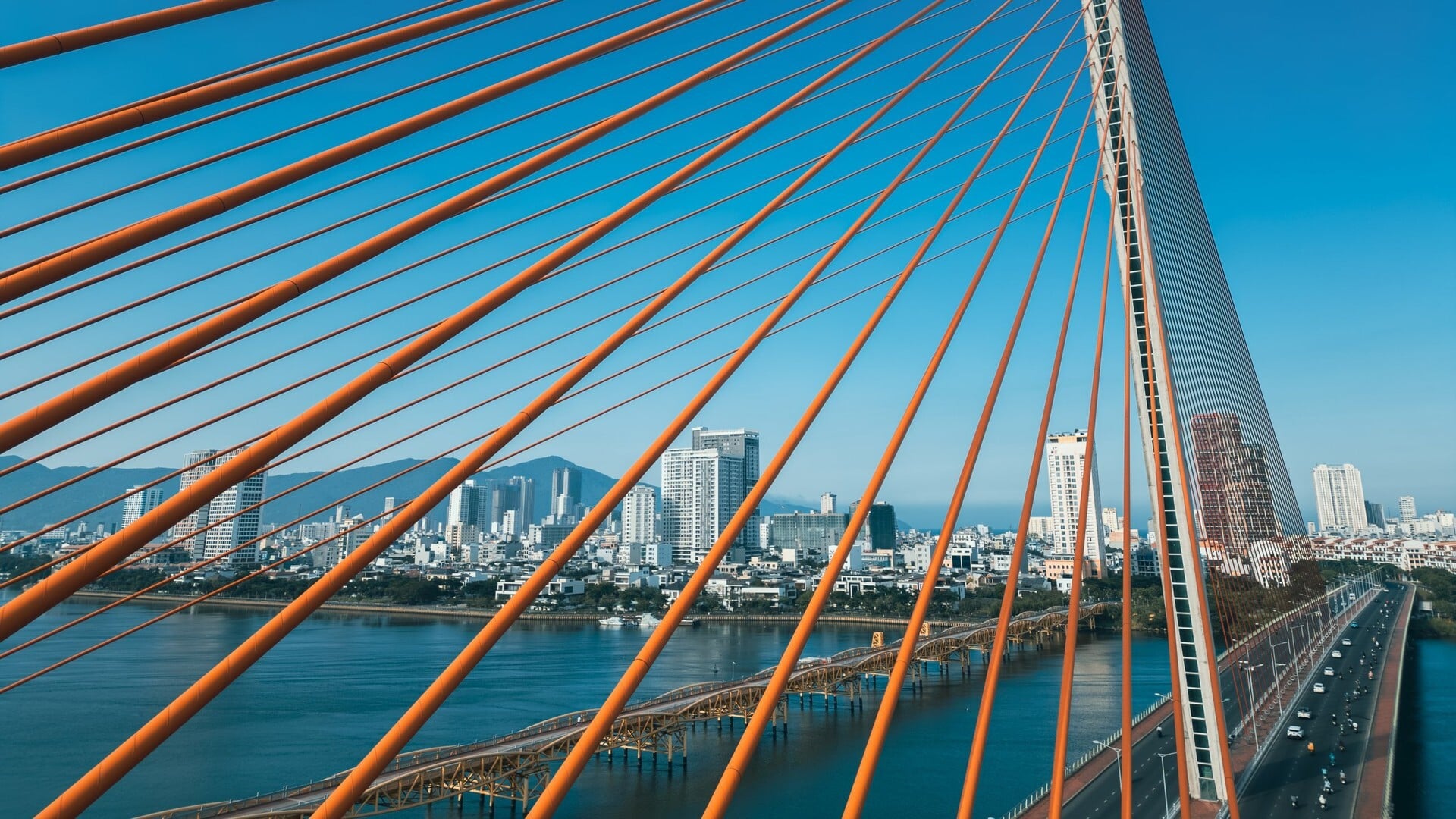 Orange cables of a cable-stayed bridge fan across a blue sky, framing a riverside city skyline with mountains behind.