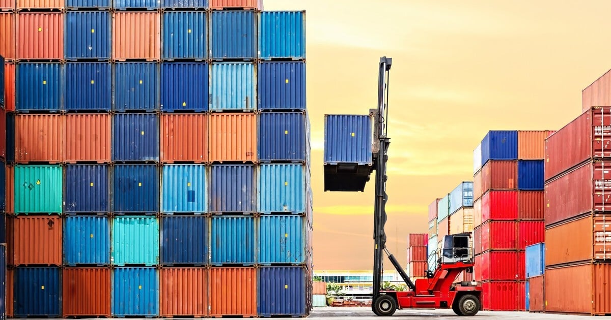 A container handler lifts a blue shipping container beside tall, organized stacks of colorful containers at a port at sunset.