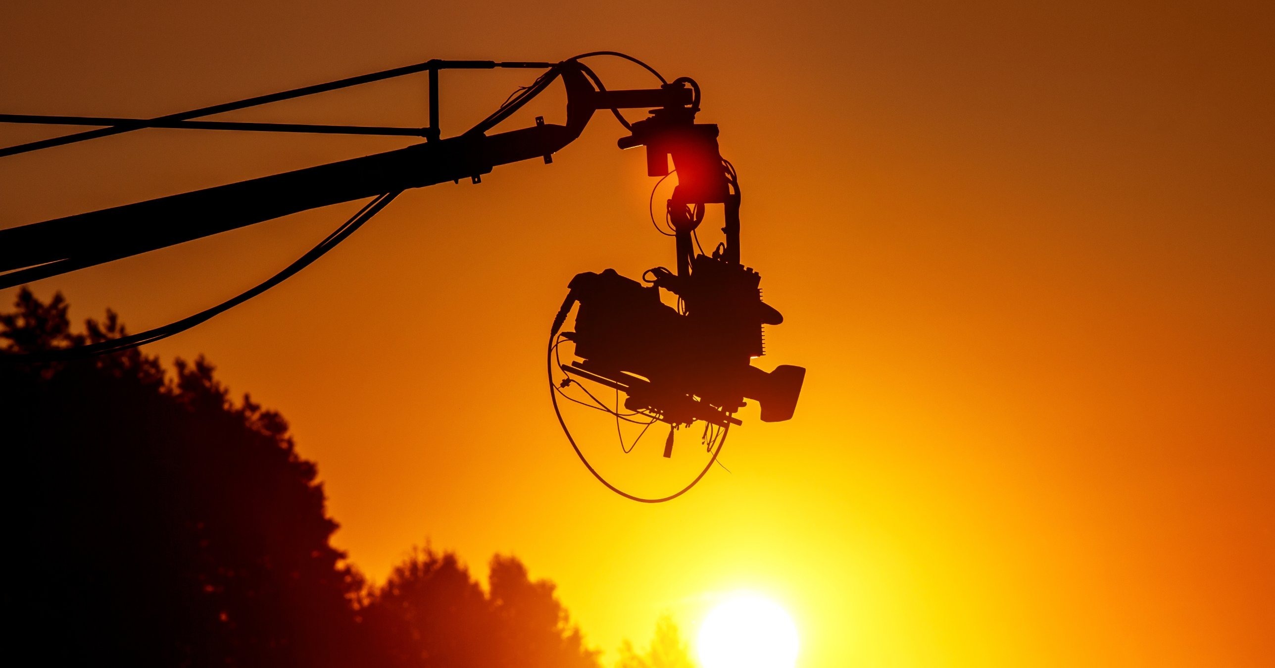 Silhouette of a camera on a crane arm against an orange sunset, with trees and the setting sun below