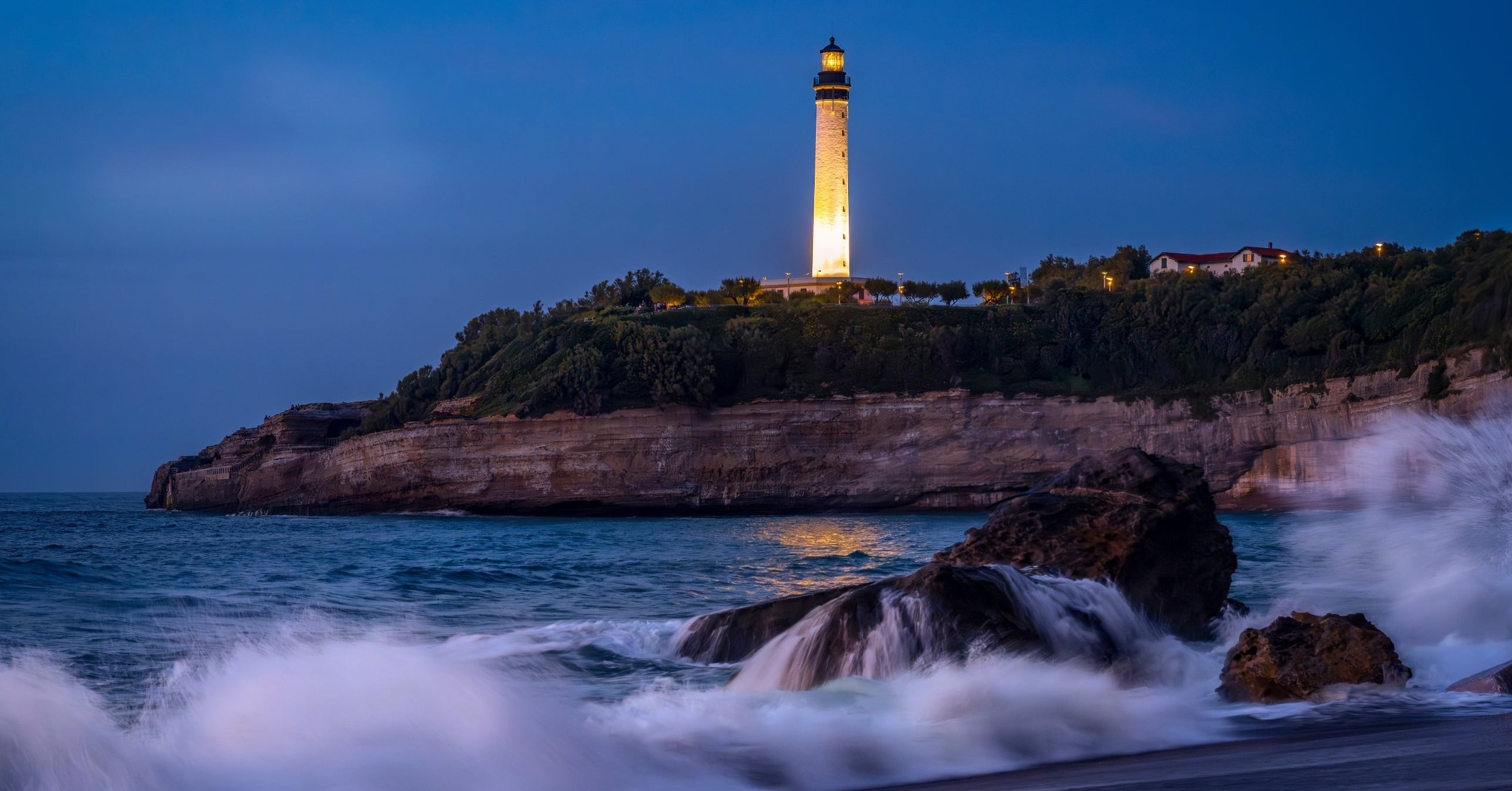 An illuminated lighthouse on a green clifftop at dusk, with waves crashing over rocks below.